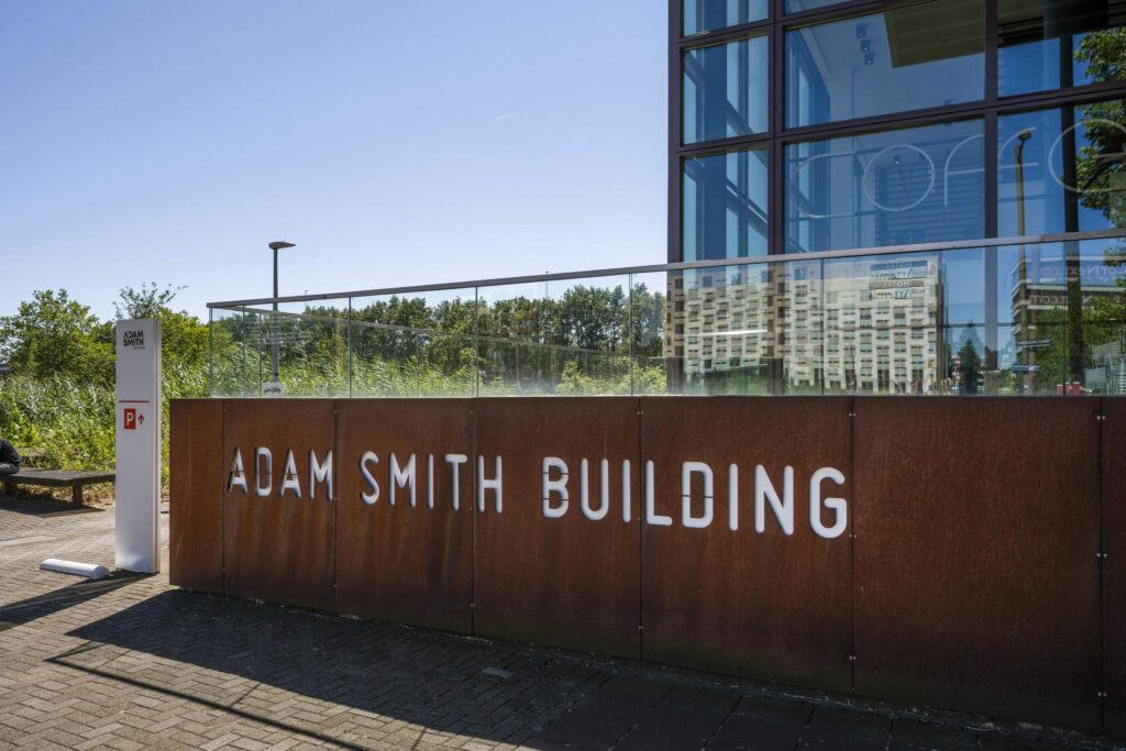 Vooraanzicht van het Adam Smith Building aan de Thomas R. Malthusstraat met moderne glazen gevel en roestbruine wand met witte belettering.